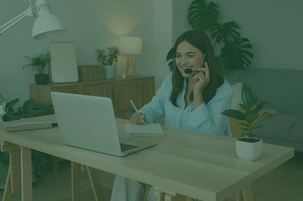 Smiling woman wearing a headset, sitting at a wooden desk with a laptop and taking notes during a video call at home.